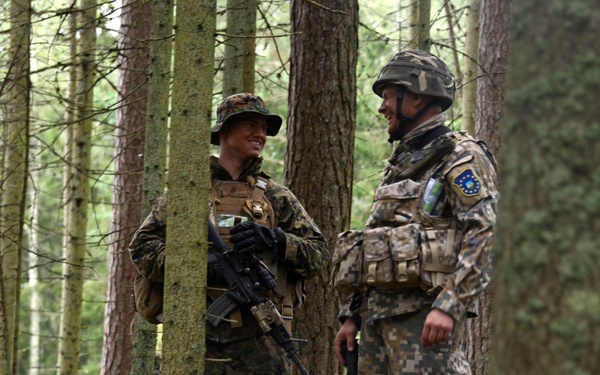 Two soldiers in different camouflage uniforms are conversing in a dense forest setting. The soldier on the left wears woodland camouflage with a boonie hat and carries an assault rifle, while the soldier on the right wears a digital/pixelated camouflage pattern with a helmet and a patch that appears to be a European Union emblem. Both appear relaxed and are smiling, suggesting a friendly interaction during a training exercise or patrol.