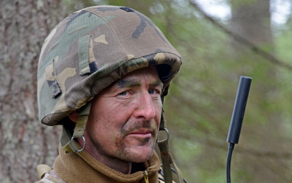 A middle-aged male soldier in a forested environment wearing mixed camouflage gear including a woodland-pattern helmet and desert digital-pattern body armor. He is equipped with tactical gear including a radio or communication device with an antenna visible on his right side. The image appears to be taken during a military exercise or field operation.
