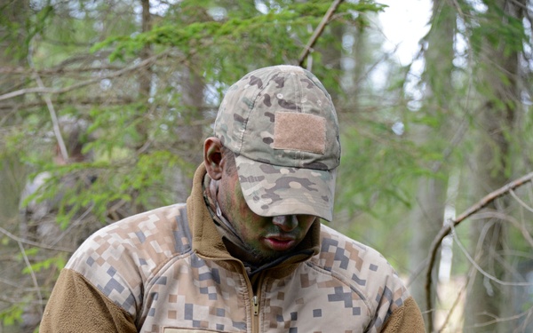 A soldier identified by name tape as 'M. MILOSS' is kneeling in a forested environment while loading ammunition into a rifle magazine. He is wearing a digital camouflage uniform with a fleece mid-layer jacket and has camouflage face paint applied. The setting appears to be a field training exercise in a boreal or northern forest environment.