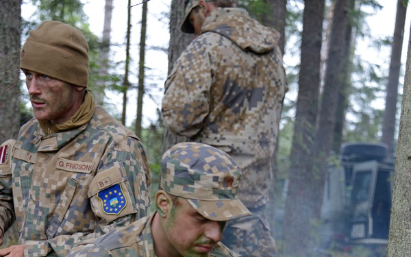 Two Latvian military soldiers in digital camouflage uniforms are resting in a forested area during what appears to be a field exercise or training operation. The foreground soldier, identified by name tape as 'LEVANDOVSKIS', is tending to a small fire or heat source, likely preparing food or water. Both soldiers wear Latvian national flag patches and EU battle group insignia on their uniforms, with green camouflage face paint applied.
