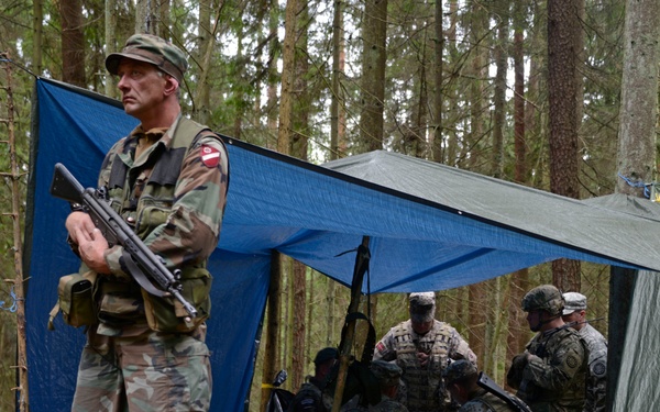 A military field exercise or operation in a forested area, showing armed soldiers in camouflage uniforms. One soldier stands guard in the foreground holding a rifle, while several others are gathered under improvised tarp shelters in the background. The setting appears to be a temporary field command post or briefing area.
