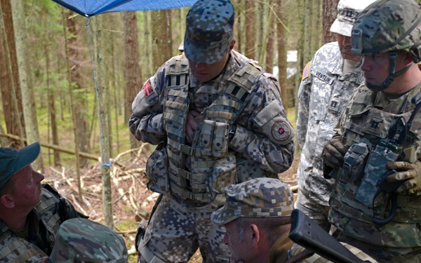 A group of multinational military personnel are gathered around a field table in a forested area, reviewing maps and documents in what appears to be a tactical planning session or military exercise. Soldiers wearing different camouflage patterns and national insignia are visible, suggesting a joint or coalition training operation. The setting includes a makeshift field command post with a tarp shelter.