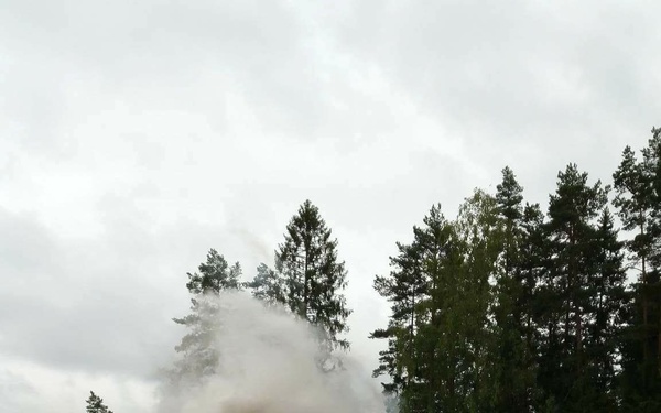 A soldier in camouflage uniform and green helmet is observing from a concealed position in dense vegetation. In the mid-distance, a large explosion or detonation is producing a significant cloud of white and grey smoke at the edge of a forested area. The setting appears to be a military training exercise in a northern European forest environment.