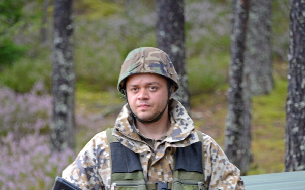 A soldier in full military gear stands in a forested environment holding a rifle. He is wearing a digital camouflage uniform and helmet, positioned next to a military-green vehicle. The background shows a sparse woodland setting with heather-like ground vegetation.