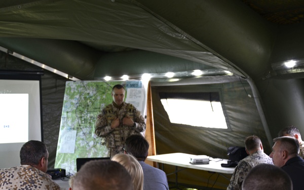A military briefing is taking place inside a large inflatable field tent. A soldier in camouflage uniform stands at the front presenting to a mixed audience of military personnel in various uniforms and at least one civilian. A projection screen and a tactical map board are visible behind the presenter.