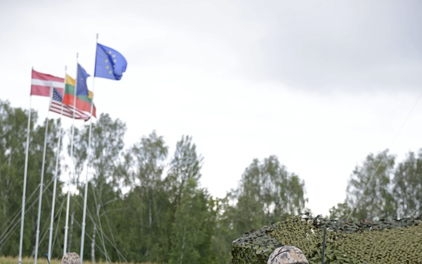 The image shows military personnel in digital camouflage uniforms conducting what appears to be a multinational military exercise or event. Multiple flags are visible in the background, including the EU flag, US flag, Latvian flag, and Lithuanian flag, suggesting a NATO or EU-affiliated joint exercise. Soldiers are armed and positioned around a camouflage-netted structure in an open field.