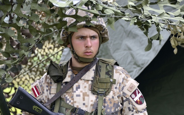 A young soldier in digital camouflage uniform stands at attention holding an assault rifle. He is positioned under a camouflage netting near a military tent, wearing full combat gear including a tactical vest and boonie hat. Shoulder patches visible on his uniform appear to include a red-and-white striped flag consistent with Latvian military insignia.