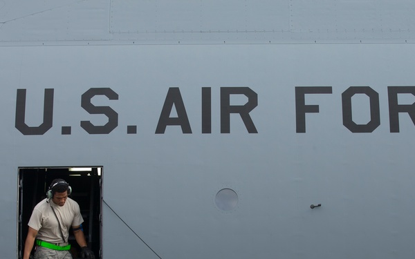 A U.S. Air Force C-5M Super Galaxy military transport aircraft, tail number 4062, is shown on the ground. A single crew member or ground technician is standing in the open side door at the top of a metal access ladder. The aircraft's fuselage is painted in standard military gray with large 'U.S. AIR FORCE' lettering and the C-5M Super Galaxy logo visible.