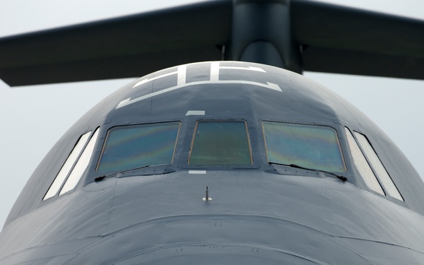 A close-up frontal view of a large military transport aircraft, photographed from ground level looking upward at the nose and cockpit section. The aircraft is painted in a flat dark gray finish typical of military aviation. The perspective emphasizes the massive, rounded fuselage and multi-pane cockpit windows.