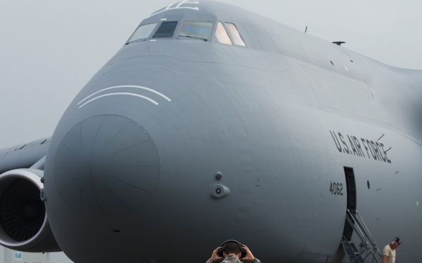 A large U.S. Air Force C-5 Galaxy military transport aircraft (tail number 4062) is parked on a flight line apron in overcast conditions. Multiple ground crew personnel in military uniforms are conducting pre-flight or post-flight servicing operations around the aircraft. The scene appears to be at a military airfield, possibly overseas based on the surrounding vegetation and infrastructure.