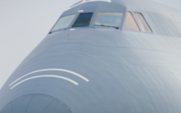 A U.S. Air Force airman in ABU (Airman Battle Uniform) camouflage stands on a flight line facing the nose of a large gray military transport aircraft, consistent with a C-130 Hercules or similar airlifter. The airman is wearing hearing protection headset and carrying a backpack, appearing to conduct a pre-flight or ground crew inspection. The overcast sky and wet tarmac suggest operations in a cool, overcast environment.