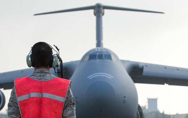 A military ground crew member in camouflage uniform and high-visibility safety vest is marshalling a large military transport aircraft on a tarmac. The aircraft, consistent with a C-5 Galaxy or similar heavy-lift transport, is taxiing directly toward the marshal. The scene appears to be at a military airfield under overcast or hazy conditions.