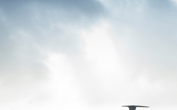 A large military or cargo transport aircraft is positioned on a tarmac apron, likely at a military airbase or airport. Several personnel in light-colored uniforms are walking across the apron in the vicinity of the aircraft. The scene is backlit with hazy, overcast sky conditions, giving the image a silhouetted, atmospheric quality.