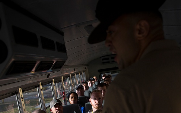 The image shows the interior of a bus with multiple passengers seated and attentively facing toward a person standing at the front. The standing individual, partially silhouetted, appears to be addressing or briefing the seated group. The passengers wear casual clothing and some have lanyards or badges visible.