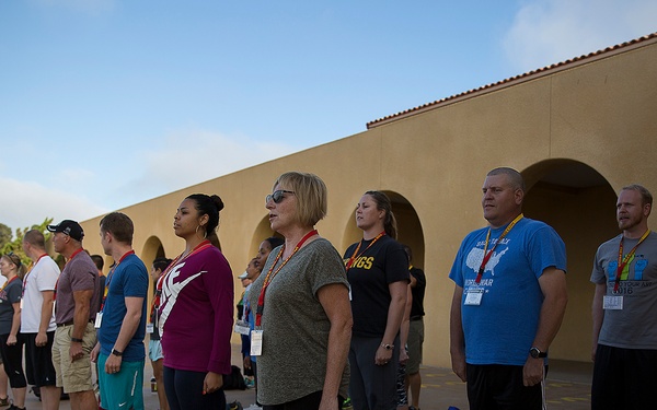 A group of civilians in athletic wear stand in formation on an outdoor paved courtyard, appearing to be at attention or receiving instruction. The setting appears to be a military base or similar institutional facility, suggested by the Spanish Colonial Revival-style building and the presence of at least one uniformed individual on the left. Participants are wearing lanyards/badges and have personal gear bags and water bottles at their feet, suggesting an organized program or event.