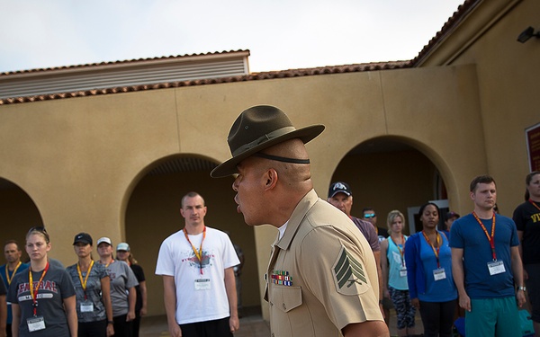 A U.S. Marine drill instructor in dress uniform with campaign cover (drill sergeant hat) addresses a group of civilians standing at attention in an outdoor courtyard. The civilians are wearing casual clothing and lanyards with badges, suggesting this is an organized orientation or educational program. The setting appears to be a military installation with a tan stucco building in the background.