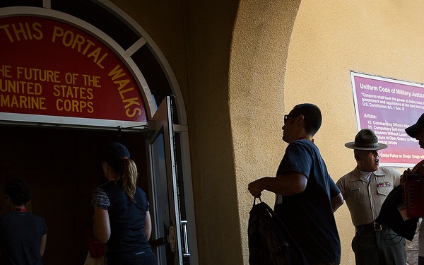 The image shows civilians and military personnel entering a building through an arched doorway bearing the inscription 'Through This Portal Walks The Future Of The United States Marine Corps.' A Marine in dress uniform with a campaign cover (drill instructor hat) is visible standing near the entrance. A sign on the wall references the Uniform Code of Military Justice, suggesting this is likely a Marine Corps recruiting or processing facility.