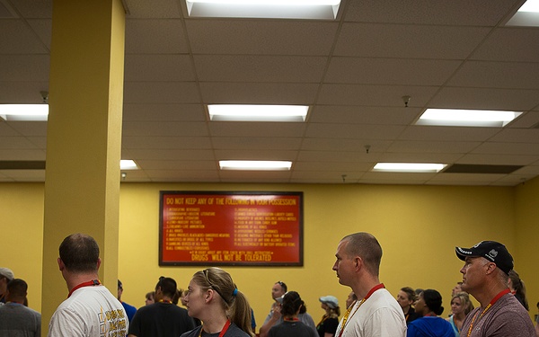 A group of people are gathered in an indoor facility with red and yellow walls, many wearing lanyards or badge credentials around their necks. They appear to be waiting in a queue or gathering area, possibly at a correctional facility, government building, or secure venue based on the visible rules/regulations sign on the wall. The space features fluorescent lighting, a drop ceiling, and red barriers or dividers in the foreground.