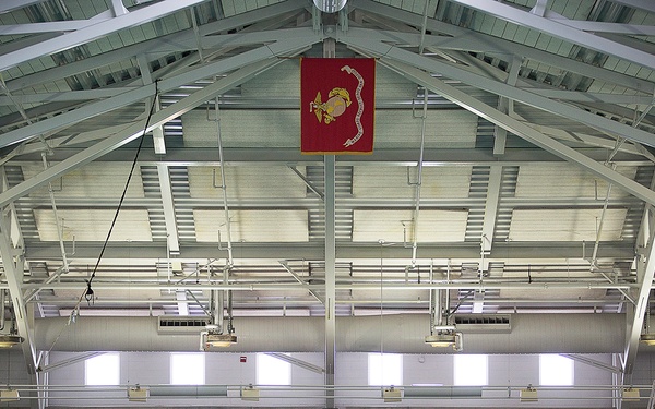 An indoor military swimming facility with a large group of spectators seated in bleachers along the far end of the pool. A red banner featuring what appears to be the United States Marine Corps emblem (Eagle, Globe, and Anchor) hangs prominently from the peaked roof. The facility features a competition-style pool with lane markers, industrial steel roof trusses, and elevated platforms on both sides.