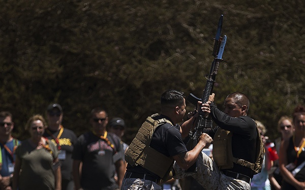 Two individuals in military camouflage uniforms and tactical vests are performing a close-quarters combat or weapons retention demonstration for a crowd of civilian spectators. One person is controlling a rifle with a blue training barrel, while the other appears to be executing a disarming or grappling technique. The event appears to be a public military demonstration or open house.