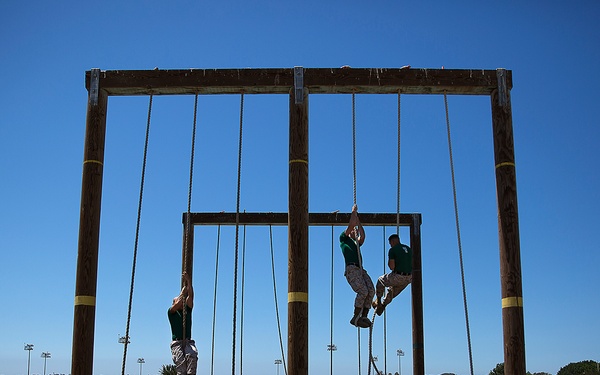 A group of military personnel and civilians observe participants climbing ropes on a large wooden obstacle course structure. The scene appears to take place at a military training facility, likely a base, under clear blue skies. Several individuals in camouflage uniforms watch as climbers ascend ropes suspended from a tall wooden frame.