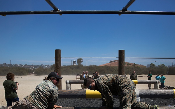 Two individuals in military camouflage uniforms are navigating a log obstacle course, likely at a military training facility. They are climbing over horizontal logs marked with yellow and black safety stripes, while observers and a photographer watch in the background. The setting appears to be an outdoor obstacle course on a sandy training ground under clear blue skies.