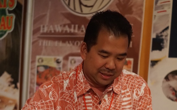 A man named Mike Yonemura, wearing a Hawaiian-print shirt and a name badge identifying him as affiliated with 'Head Seafood,' is serving food at what appears to be the 2016 Hawaii Exhibition. He is using tongs to plate what appears to be poke (Hawaiian raw fish dish) with various toppings into a bowl. Several bowls of prepared seafood dishes are arranged on a tray in front of him.