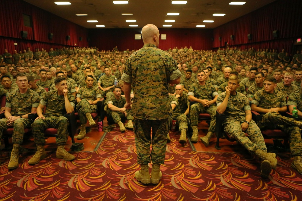 A senior military officer in USMC MARPAT camouflage uniform stands at the front of a large auditorium addressing hundreds of seated Marines. The venue appears to be a base theater or large briefing hall with red curtains, patterned carpet, and rows of theater-style seating. The audience is attentive and filling the hall to near capacity.