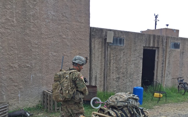 A soldier in full combat gear and camouflage uniform operates or guides a tracked robotic ground vehicle loaded with military equipment and supply crates. The scene takes place in a compound with mud-brick or concrete structures typical of rural Central or South Asian architecture. Several bicycles, a blue water barrel, and wooden crates are visible in the background.