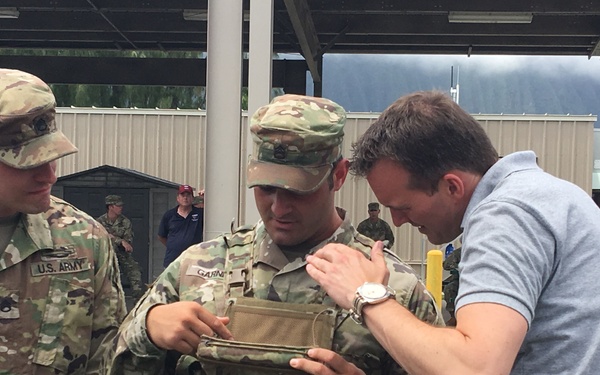 Two U.S. Army soldiers in OCP (Operational Camouflage Pattern) uniforms are interacting with a civilian man in a gray polo shirt at what appears to be a military installation. The civilian is examining or adjusting a tactical plate carrier vest worn by one of the soldiers. The setting appears to be an outdoor covered area at a military base, with mountains visible in the background.