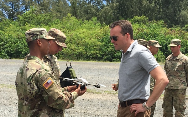 A group of U.S. Army soldiers in OCP (Operational Camouflage Pattern) uniforms are gathered on a gravel area surrounded by tropical vegetation. A soldier is demonstrating a small unmanned aerial vehicle (UAV/drone) to a civilian man in a gray polo shirt and khaki pants. The setting appears to be an outdoor military demonstration or briefing, likely in a tropical location such as Hawaii.