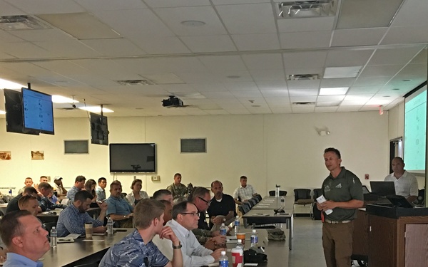 A formal briefing or conference is taking place in a large meeting room, likely on a military installation, as indicated by the red general officer flag visible on the right side. A male presenter stands at a podium delivering a presentation to approximately 30-40 attendees seated at rows of tables. The room is equipped with multiple display screens, a ceiling-mounted projector, and a projection screen showing a green-tinted slide.