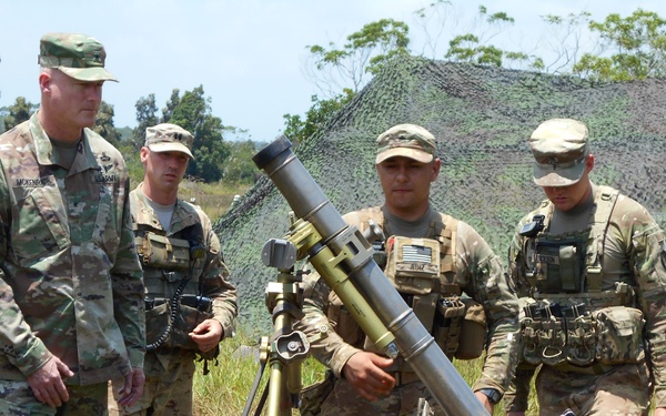 Four U.S. Army soldiers in MultiCam/OCP camouflage uniforms are gathered around a mortar system mounted on or near a military vehicle in an outdoor field setting. One soldier on the left appears to be a senior officer based on rank insignia, and a name tape reading 'MCKENZIE' or similar is partially visible. The group appears to be conducting a demonstration or inspection of the mortar weapon system.