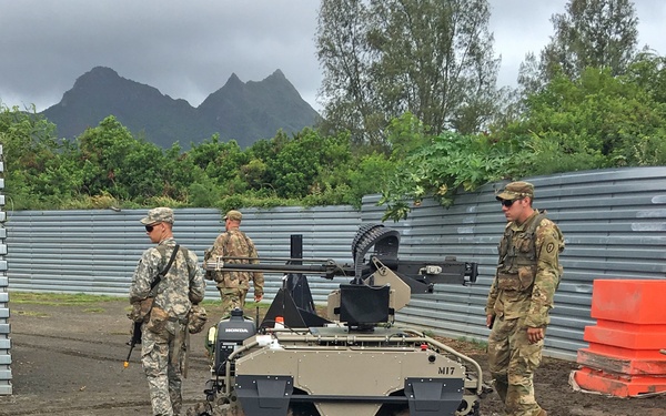Three military personnel in camouflage uniforms are observing a small unmanned ground vehicle (UGV) on a gravel surface. The tracked robotic platform, marked 'M17' and equipped with a weapon system mount and a Honda engine, appears to be undergoing evaluation or demonstration. The setting features a tropical mountainous backdrop consistent with Hawaii or a similar Pacific location.
