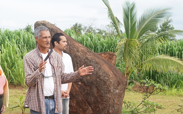 Four people are gathered outdoors near a large volcanic or basalt boulder in what appears to be a Hawaiian setting, indicated by the red soil, palm tree, and lei worn by one individual. A man in a plaid shirt appears to be explaining or presenting something about the site, gesturing toward the boulder and surrounding area. The background features dense green sugarcane or similar tall grass crops.