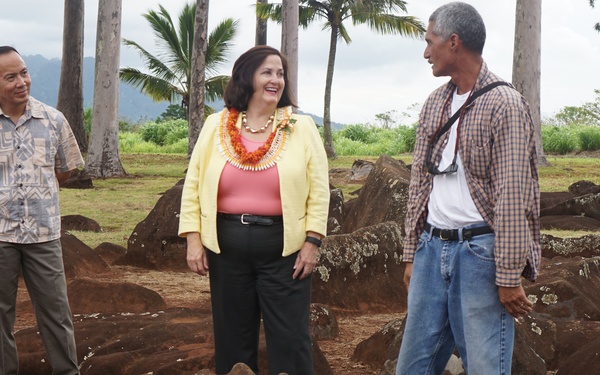 Three adults are standing among large volcanic or basalt rocks in what appears to be a Hawaiian archaeological or cultural site, likely a heiau (traditional Hawaiian temple). The central figure, a woman wearing a yellow jacket and orange lei, appears to be receiving a tour or official visit. The setting features red soil, scattered large stones, palm trees, and mountainous terrain in the background.