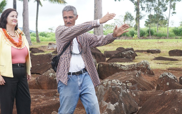 Two individuals stand among a field of large, reddish-brown volcanic rocks scattered across red soil, surrounded by tropical vegetation including palm trees. A man in a plaid shirt and jeans gestures outward, appearing to explain or point out features of the site to a woman wearing a yellow blazer and a traditional Hawaiian lei. The setting appears to be a Hawaiian archaeological or cultural heritage site.