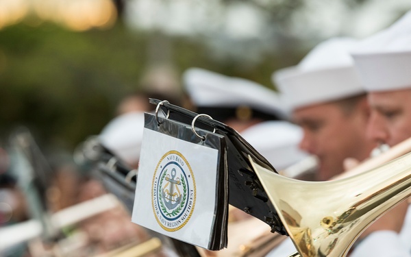A U.S. Navy band is performing outdoors, with multiple sailors in white dress uniforms playing trombones. A music stand attachment on one of the trombones displays a seal reading 'Navy Band' with an anchor emblem, consistent with a Navy Region Southwest insignia. The image captures a close-up perspective of the brass instruments and musicians during what appears to be a parade or ceremonial performance.