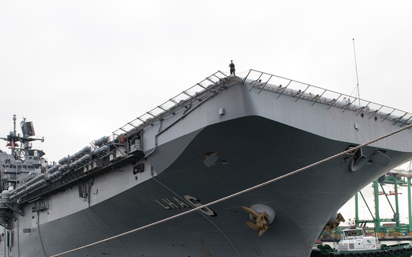 A large U.S. Navy amphibious assault ship is docked at a commercial or naval port, viewed from a low angle at the bow. The hull markings appear to read 'LHA-6', suggesting this is the USS America or a similar Wasp/America-class vessel. A tugboat is visible alongside the starboard side, and green container cranes are visible in the background.
