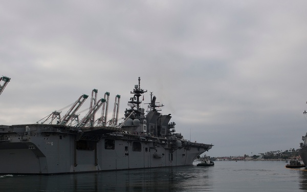 A large U.S. Navy amphibious assault ship is maneuvering in a harbor, assisted by tugboats, while a destroyer is moored at the pier to the right. The scene appears to be at a naval installation, likely on the U.S. West Coast, with commercial container cranes visible in the background. Weather conditions are overcast with calm water providing clear reflections of the vessels.