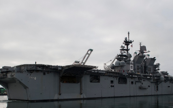 A large US Navy amphibious assault ship bearing the hull designation 'LHA 6' is photographed in a harbor under overcast skies. The vessel is a Wasp-class or America-class amphibious assault ship, identifiable by its flat flight deck and island superstructure on the starboard side. The ship appears to be maneuvering near a commercial port facility, assisted by tugboats.