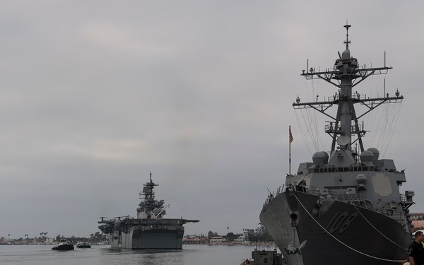 A naval pier scene showing a large US Navy guided-missile destroyer moored at a dock, with what appears to be an amphibious assault ship visible in the background waterway. Several personnel in dark uniforms stand on the pier alongside a white pickup truck. The scene is set under overcast skies with port infrastructure visible in the background.