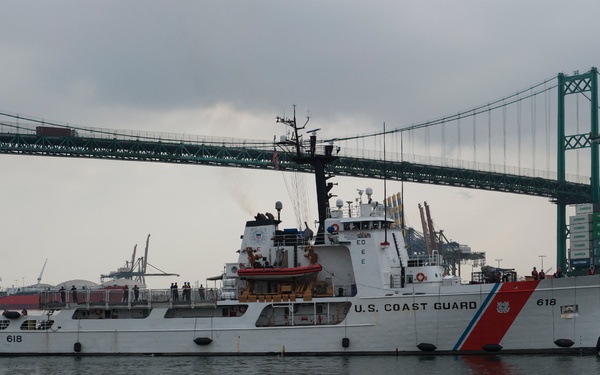 A U.S. Coast Guard vessel numbered 618 is operating in a busy harbor beneath a large suspension bridge. The white cutter displays the distinctive Coast Guard diagonal red and blue stripe, with personnel visible on deck. A large container ship and industrial port infrastructure are visible in the background.