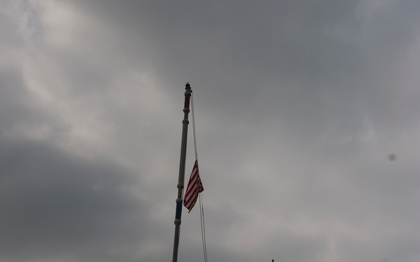 Two personnel are standing on the bow of a dark-hulled naval or military vessel, raising or handling an American flag on a tall mast. The scene is set at a port or harbor with large green container cranes visible in the background. The overcast, stormy sky creates a dramatic, low-light atmosphere.