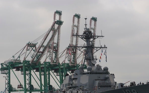 A U.S. Navy guided-missile destroyer bearing hull number 108 is being maneuvered in a commercial port, assisted by FOSS Maritime tugboats. Large green container cranes, identifiable as Evergreen terminal equipment, dominate the background. The overcast sky and calm harbor waters suggest a routine port arrival or departure operation.