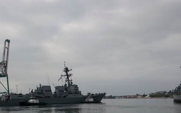 A naval harbor scene showing a U.S. Navy Arleigh Burke-class guided-missile destroyer being maneuvered into port with the assistance of tugboats. To the right, an older warship — likely a museum or reserve vessel — is moored at the dock. Large green container cranes are visible on the left side of the harbor, indicating a commercial port facility.