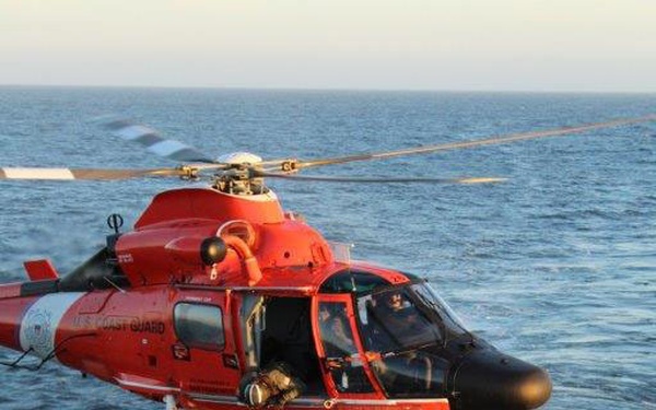 A U.S. Coast Guard helicopter (marked 'U.S. COAST GUARD') in orange-red livery hovers over the flight deck of a vessel at sea. Ground crew personnel on the deck are managing a hoist cable connected to the helicopter, while a flight deck director in a yellow vest and helmet signals the aircraft. The operation appears to be a hoist or cargo transfer exercise conducted over open ocean.