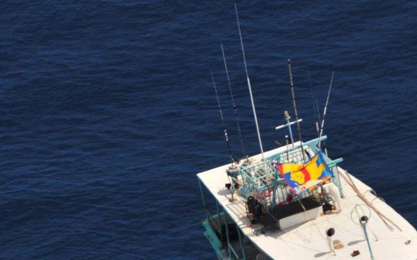 An aerial photograph shows a small orange rigid inflatable boat (RIB) approaching a larger white fishing or patrol vessel on open blue water. The RIB appears to carry several personnel, suggesting a maritime interdiction, boarding operation, or rescue scenario. The larger vessel has multiple fishing rods/antennas, a teal/turquoise hull trim, and a colorful flag displayed on its superstructure.