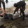 A military training or medical exercise scenario is depicted outdoors on a wet concrete surface. A soldier in camouflage uniform and black gloves is attending to a civilian-clothed person lying on the ground, likely simulating casualty care or a search procedure. At least one additional uniformed individual is visible in the background.