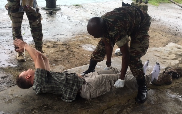A military training or medical exercise scenario is depicted outdoors on a wet concrete surface. A soldier in camouflage uniform and black gloves is attending to a civilian-clothed person lying on the ground, likely simulating casualty care or a search procedure. At least one additional uniformed individual is visible in the background.